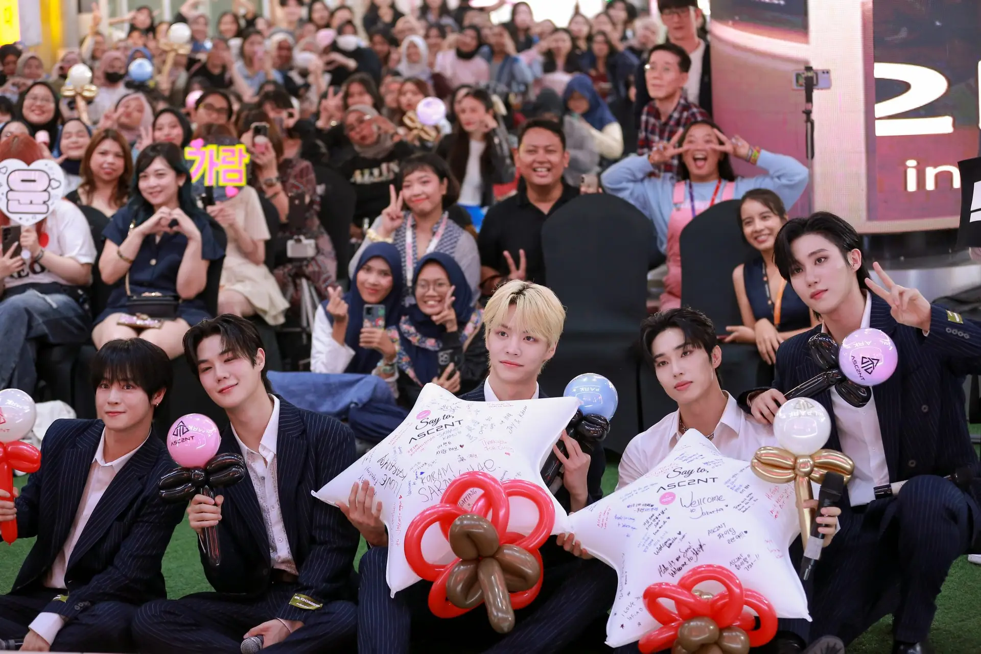 Asc2nt boy group posing with fans during a fan meeting in Lotte Mall Jakarta, Indonesia. The members are seated in the front holding balloons and decorated star-shaped pillows, while a large crowd of excited fans smiles and makes hand gestures in the background.