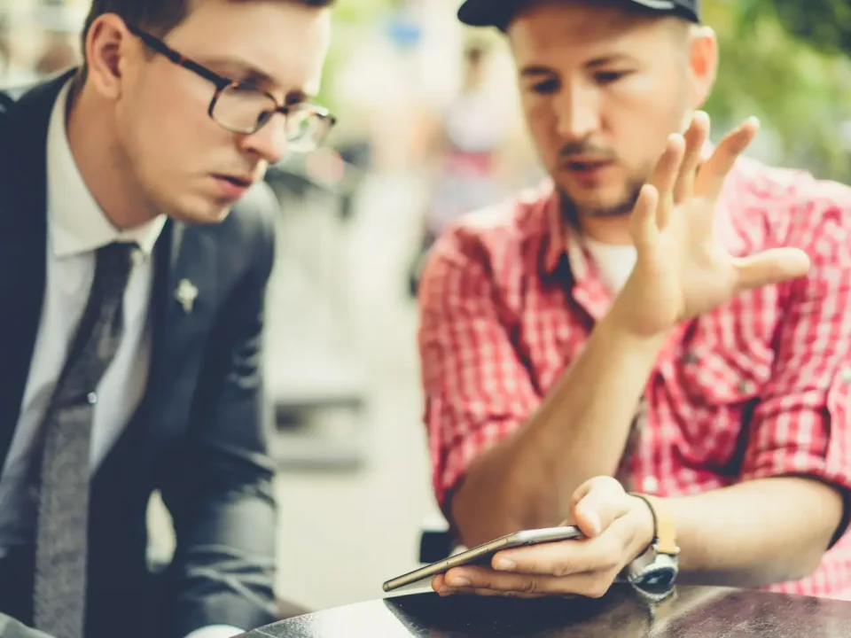 Dua orang pria sedang berdiskusi serius sambil menunjuk layar smartphone, mengilustrasikan proses analisis sentimen dan percakapan audiens dalam strategi social listening.