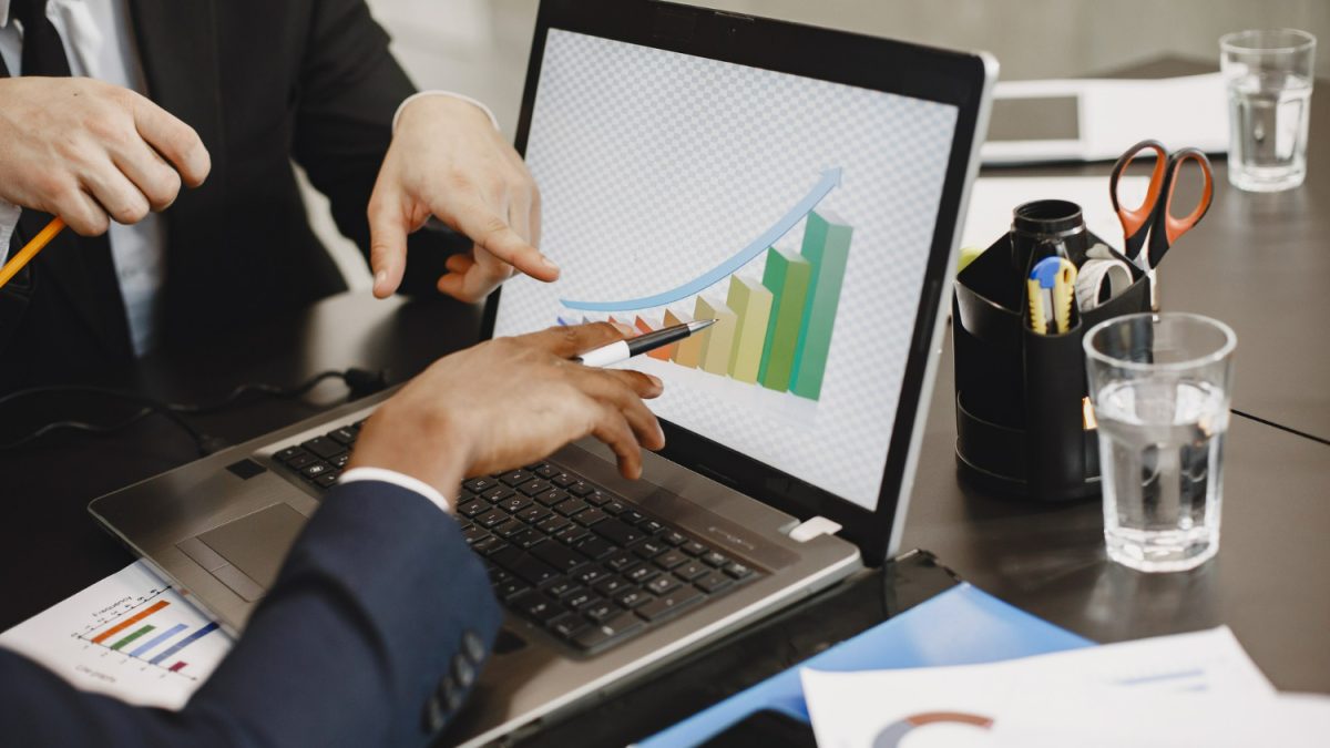 Dua pebisnis menganalisis grafik pertumbuhan di laptop, mendiskusikan angka dan strategi untuk mengukur ROI digital marketing.